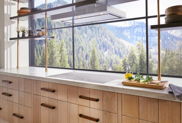 A kitchen with blonde wood cabinets, white marble counters, and an induction stove facing out to a view of a mountainside covered with pine trees.