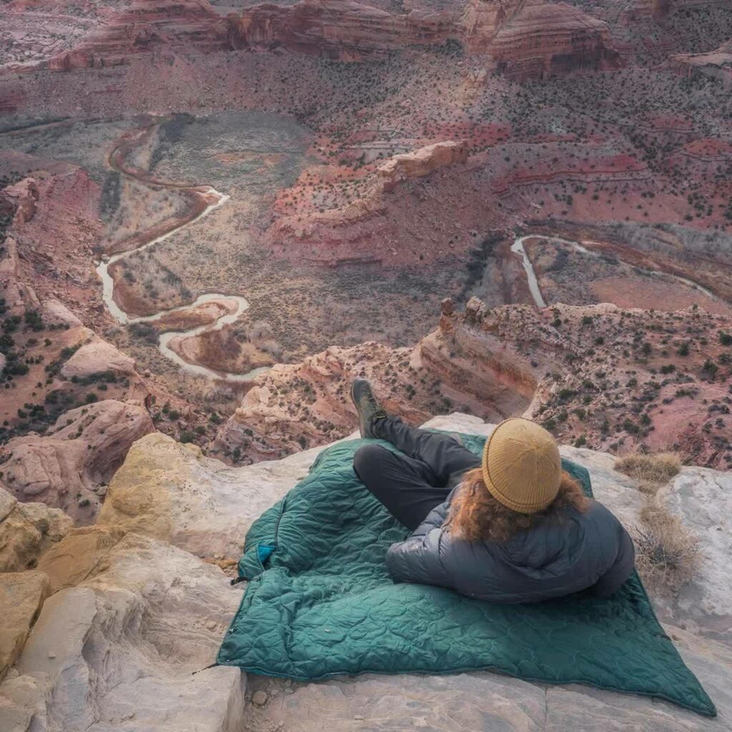 A person with curly red hair is lying down on a green blanket on a mountainside overlooking a dramatic rocky red valley.